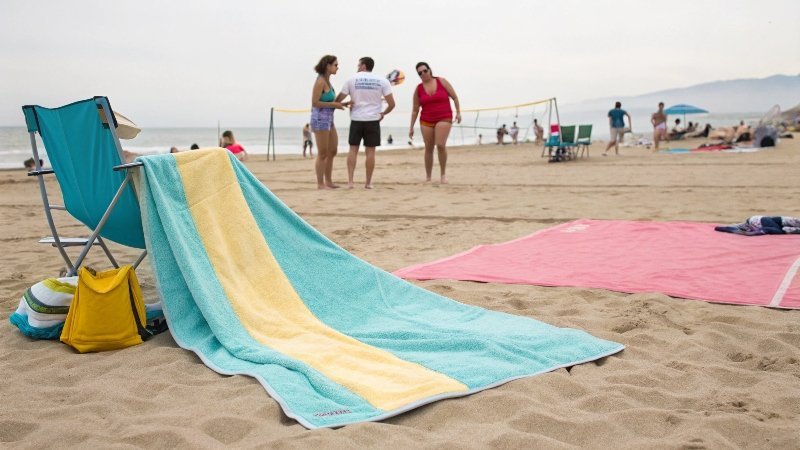 Osoba relaksująca się na dużym ręczniku plażowym na piasku, z książką i okularami przeciwsłonecznymi.